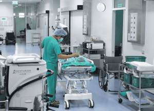 A medical staff in scrubs and a mask assisting a patient in a hospital room 